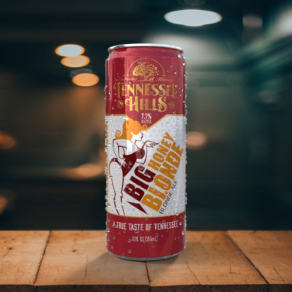 A can of Tennessee Hills Big Honey Blonde beer with a graphic design and text indicating the brand and product name, placed on a wooden surface with a blurred background.